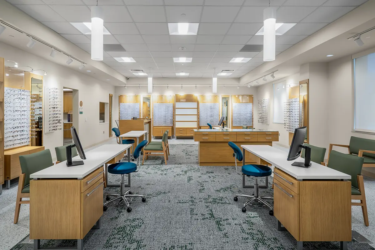 Modern optician's office with wooden desks and blue chairs, featuring bright lighting and walls lined with display shelves of eyeglasses. Calm, professional ambiance.