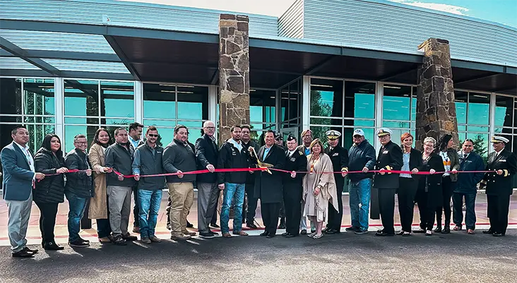 A diverse group of people stands in front of a modern building for a ribbon-cutting ceremony. They hold a long red ribbon, smiling in celebration.