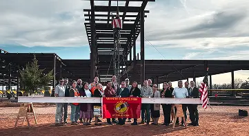 A group of people stands in front of a steel structure at a construction site, holding a white beam with a red banner. The mood is celebratory.