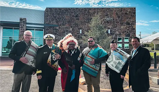 A group of six people stand smiling in front of a stone building, each holding a colorful patterned blanket. One person wears a feathered headdress. The sky is clear and sunny.