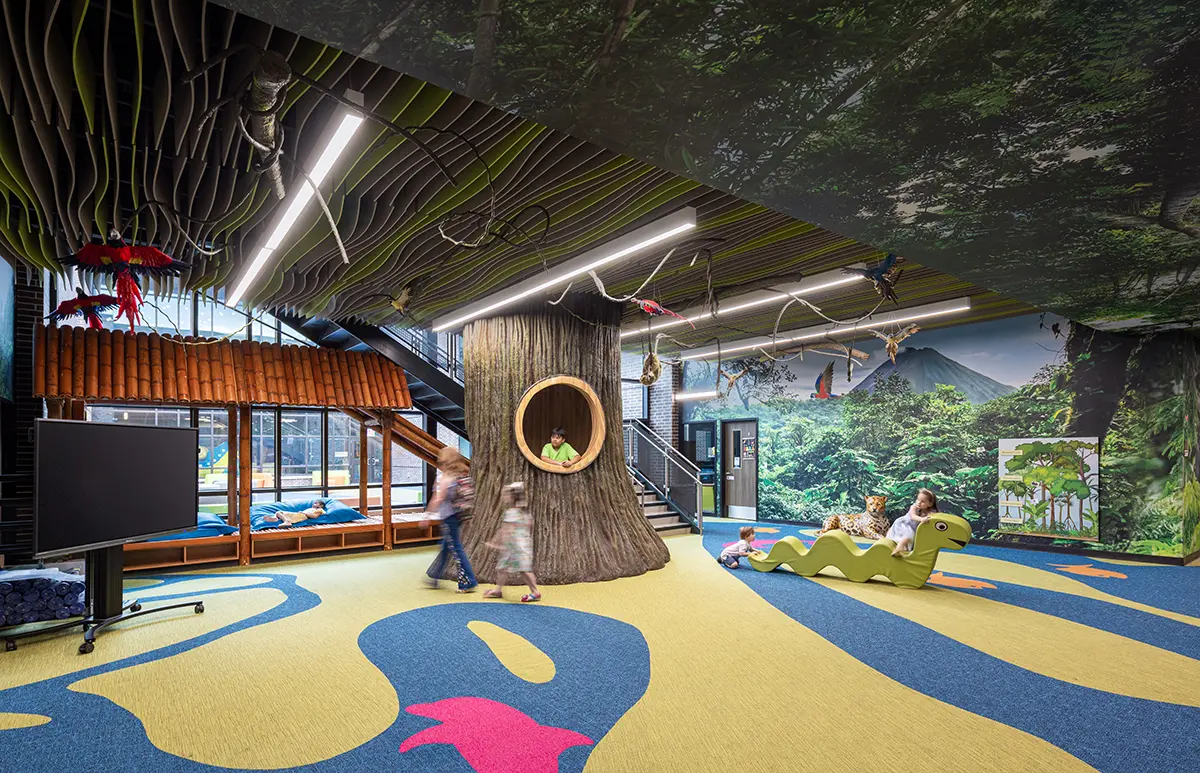Children’s play area with a jungle theme. Features include a large tree with a circular window, forest-patterned walls, and kids on a green seesaw.