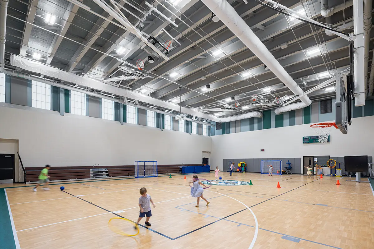 Children play in a bright gymnasium with a wooden floor and high ceiling. They are using colorful cones and hula hoops amidst a lively atmosphere.