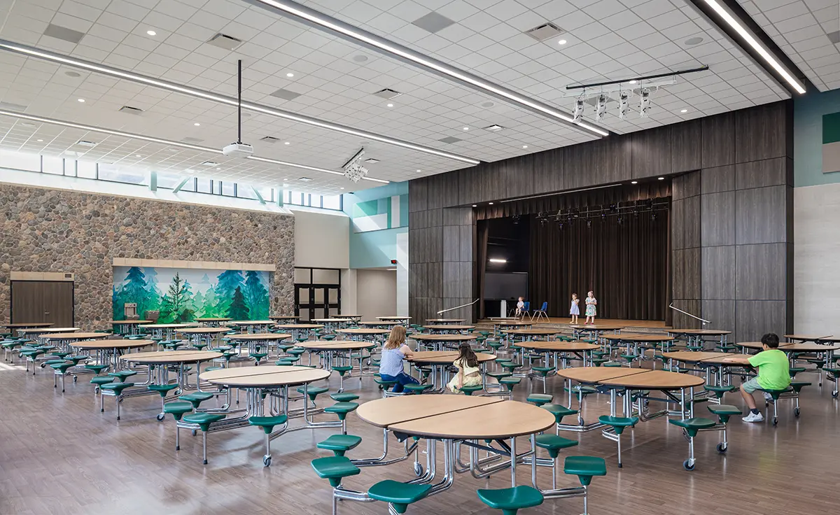 Spacious cafeteria with round tables, green seats, and a stone accent wall featuring a forest mural. A stage at the back with a few people on it. Bright, airy atmosphere.