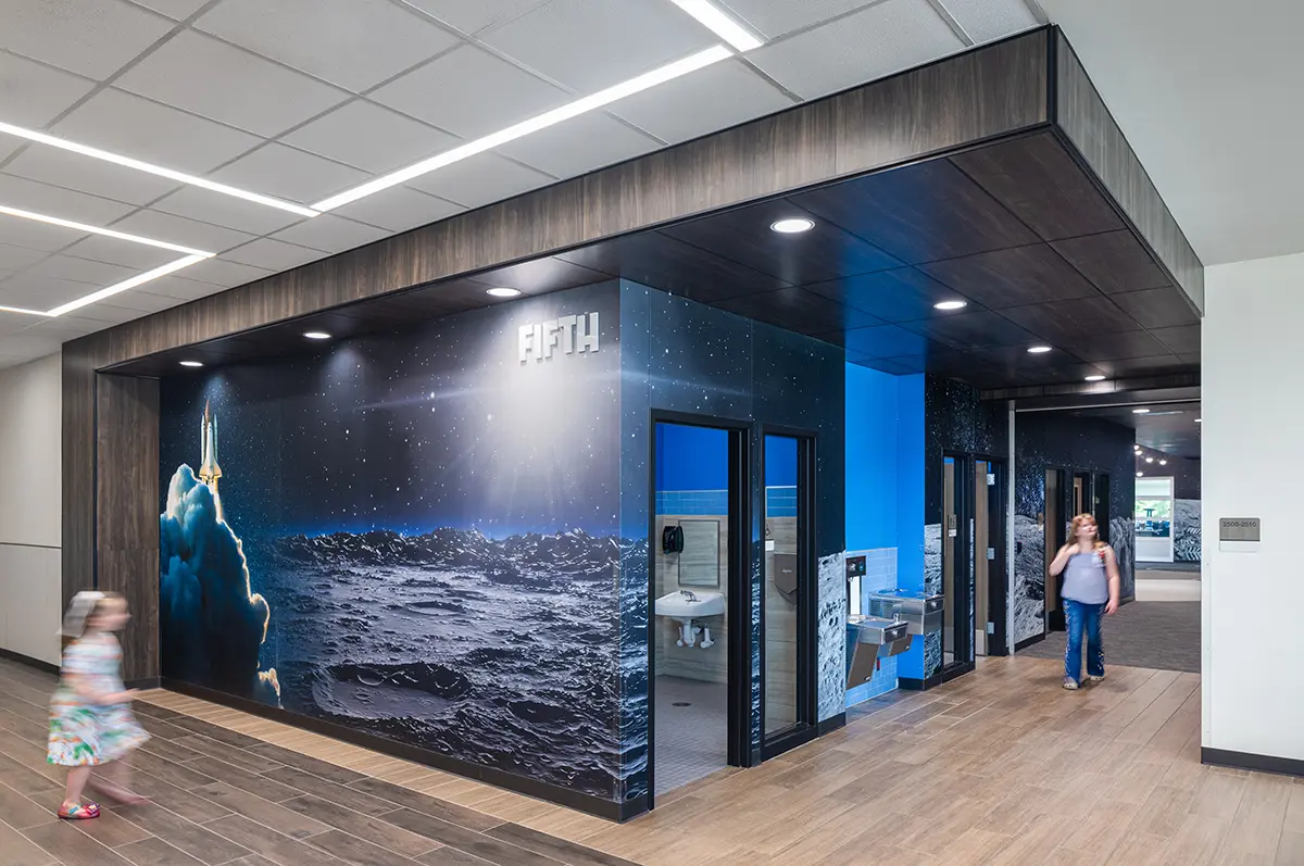 Hallway with a space-themed mural depicting a rocket launch and moon surface. Sign reads “FIFTH.” A child and adult walk nearby, creating a lively atmosphere.