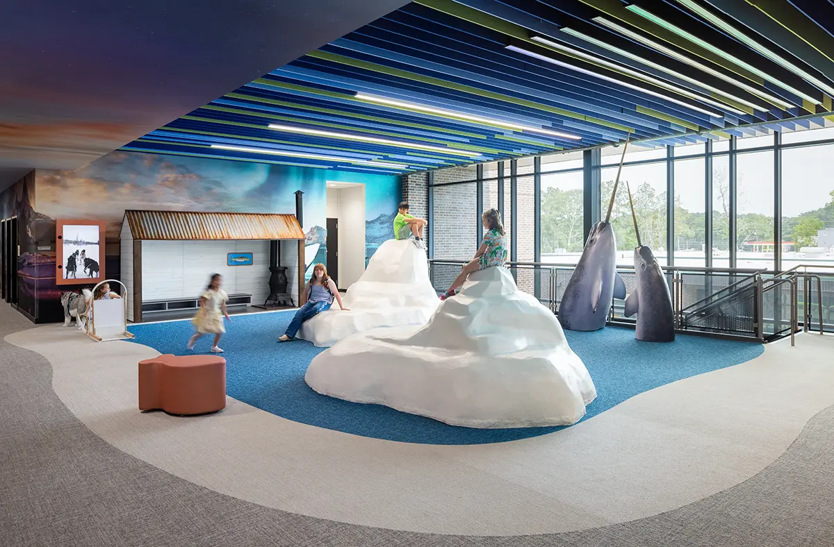 A modern play area with iceberg-themed climbing structures, children playing, and a penguin mural. The vibrant ceiling and large windows add brightness.
