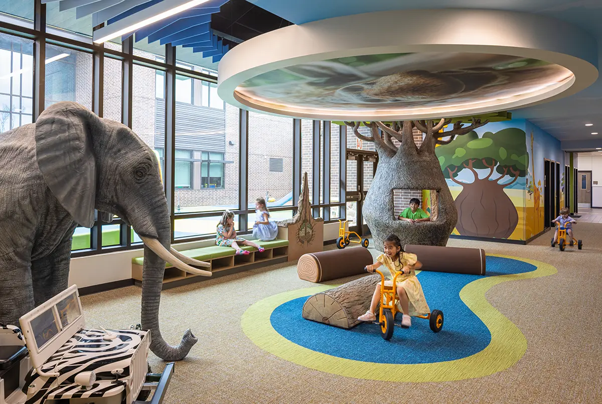 Children's playroom with safari theme; features large elephant, tree with nest, kids on tricycles, vibrant colors, playful atmosphere.
