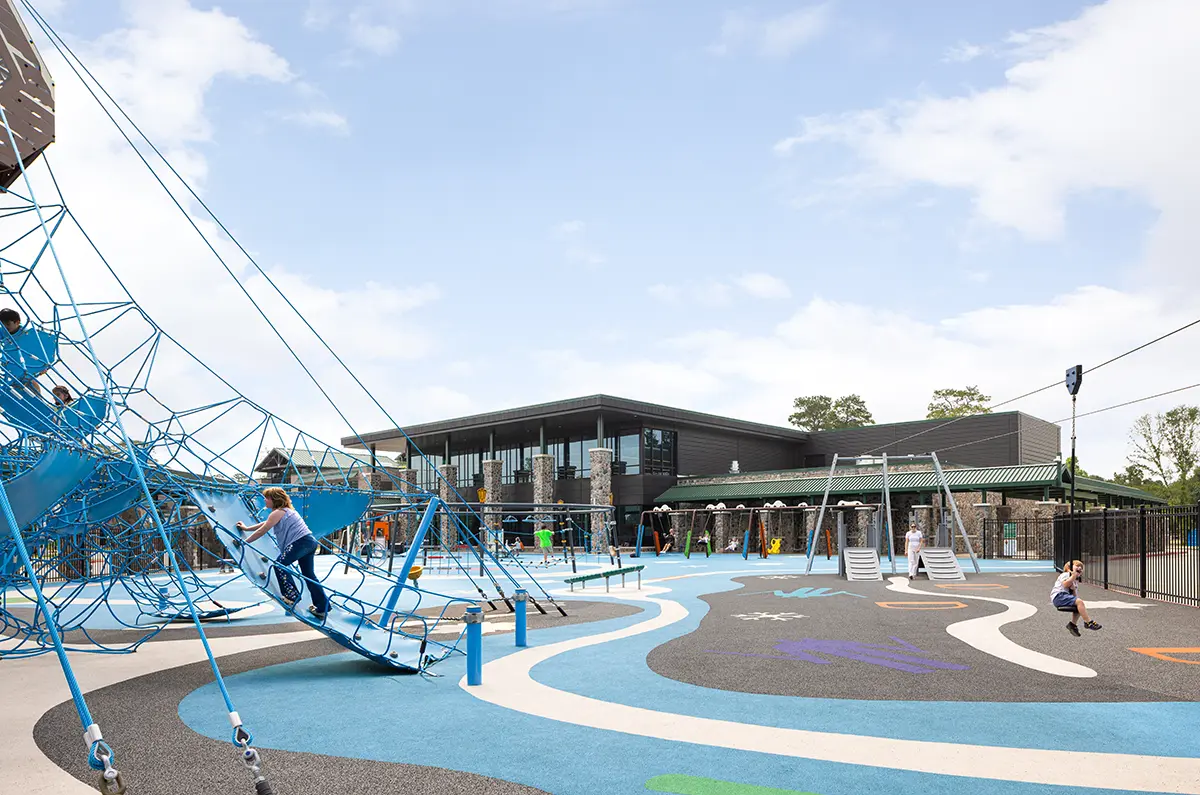 A modern playground features a blue rope climbing structure and swings. Children play, creating a lively and energetic atmosphere under a partly cloudy sky.