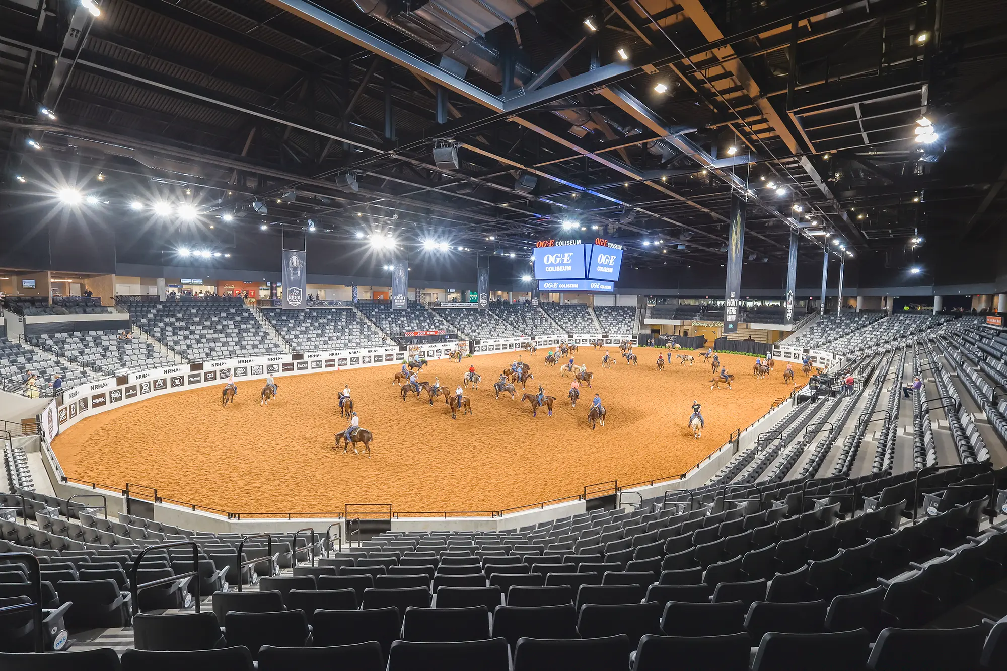 Indoor arena with a sandy floor, hosting equestrian activities. Riders and horses practice under bright lights, surrounded by empty tiered seating. Calm atmosphere.