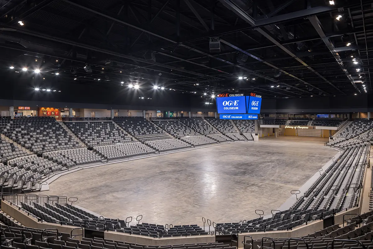 A large empty indoor arena with tiered rows of beige and gray seats. Overhead lights illuminate a central scoreboard displaying “OGE Coliseum” in blue.