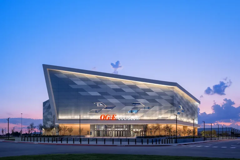 Modern arena at dusk with an angular design and illuminated entrance. The sky transitions from pink to blue, creating a vibrant, dynamic atmosphere.