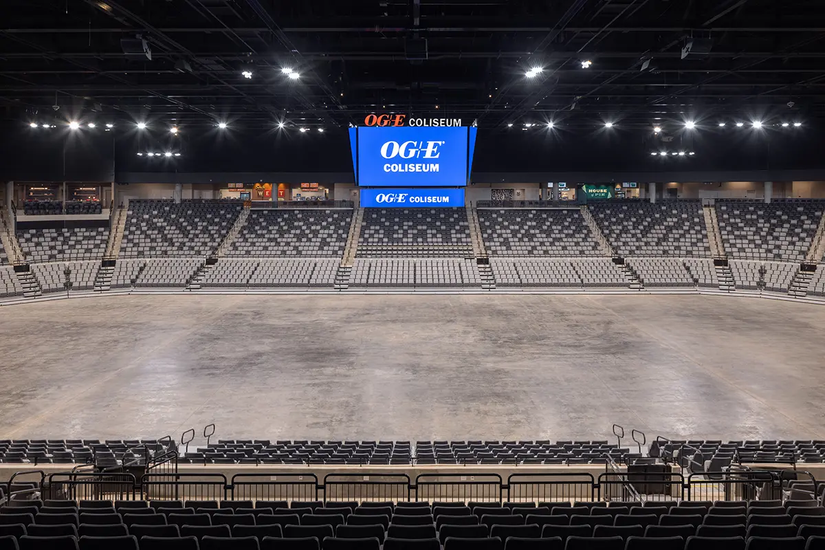 Empty indoor arena, OGE Coliseum. Gray seating surrounds a central, bare floor. A large screen displays the venue name. The atmosphere is quiet and spacious.