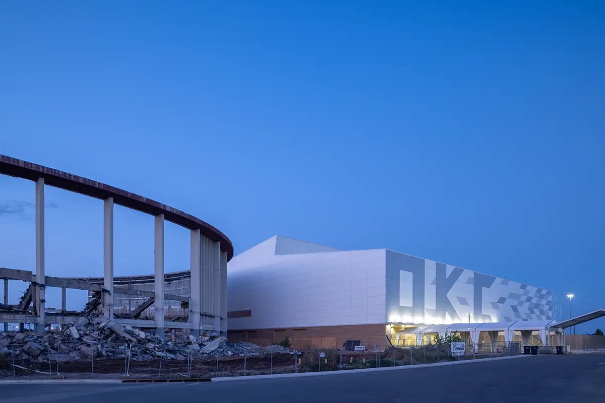 A modern, white building with "OKC" in large letters is illuminated in the twilight. Adjacent is a partially demolished structure, contrasting old and new.