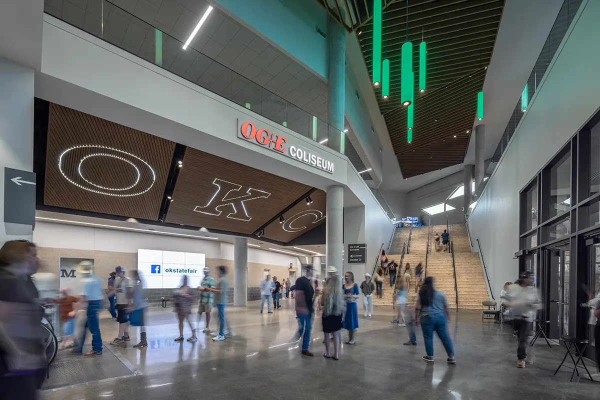 Modern coliseum interior with "OKC" sign above, bustling with people. Green vertical lights hang from the ceiling; a staircase is in the background.