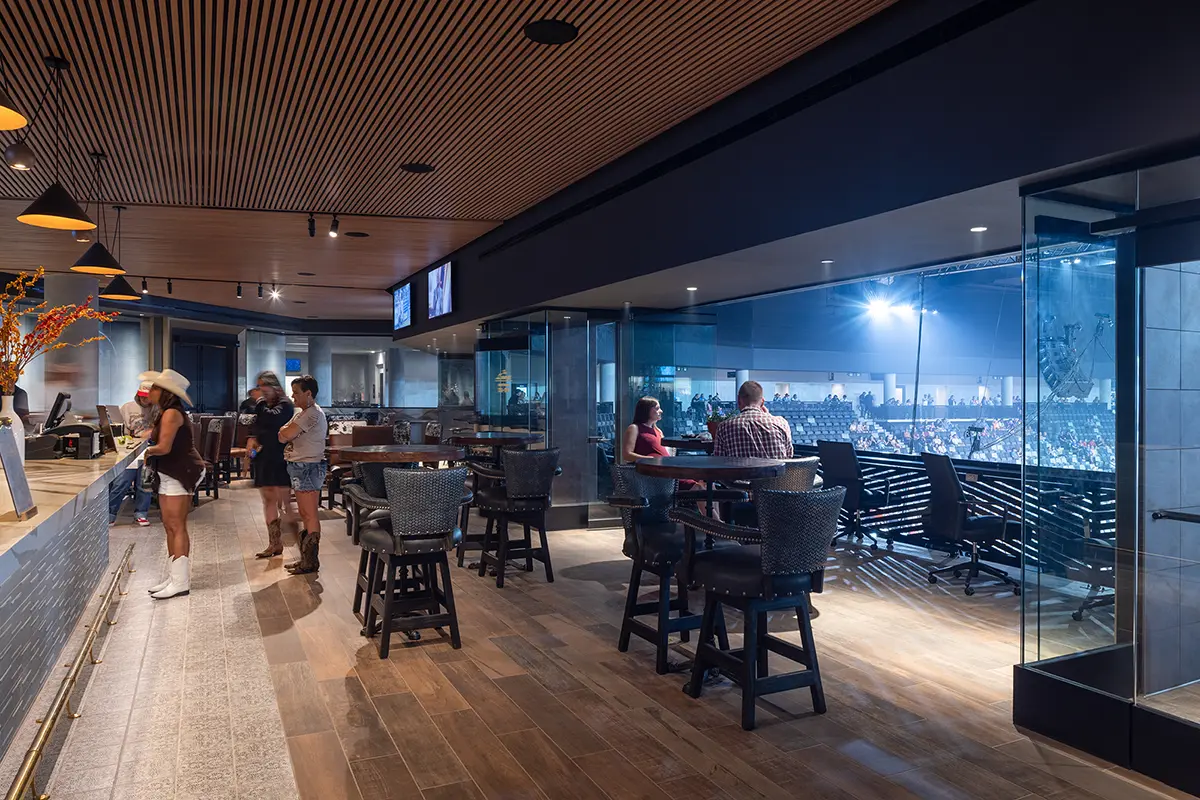 Spacious lounge with wooden floors and modern decor, features people at a bar counter. Large glass windows reveal a bright, busy arena below.