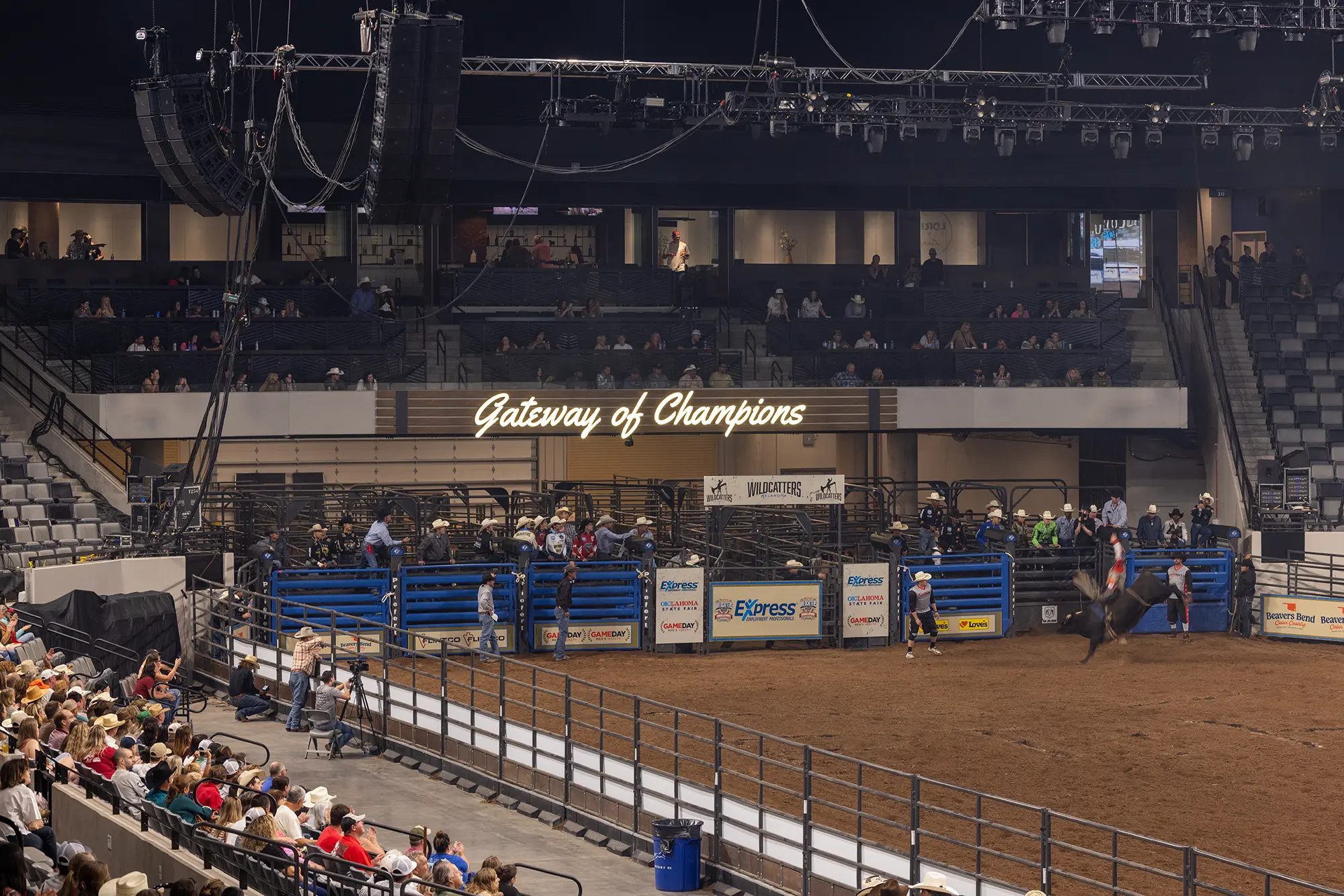 Indoor rodeo arena with a horse and rider under a sign reading "Gateway of Champions". A crowd watches from stands, creating an energetic atmosphere.