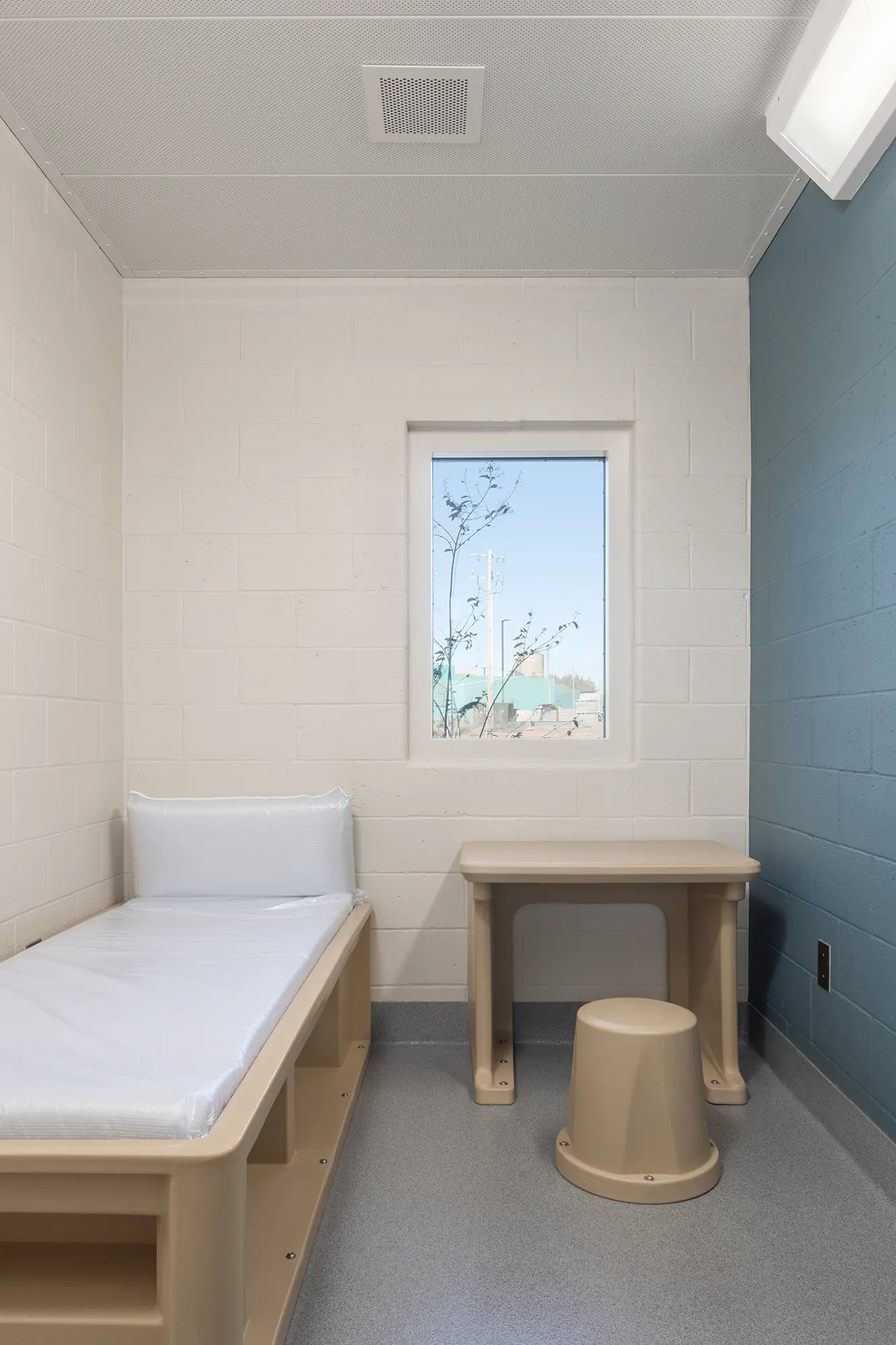 Minimalist room with a light beige bed, small table, and stool. A window offers an outdoor view. Walls are white and blue, conveying a clean, sparse feel.