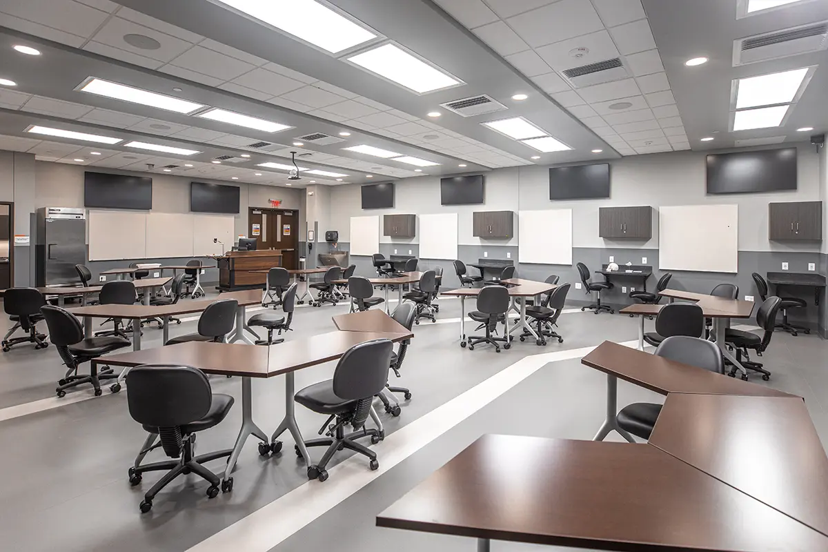 A modern, empty classroom with triangular wooden desks and black chairs arranged in clusters. Whiteboards and screens line the walls under bright lighting.
