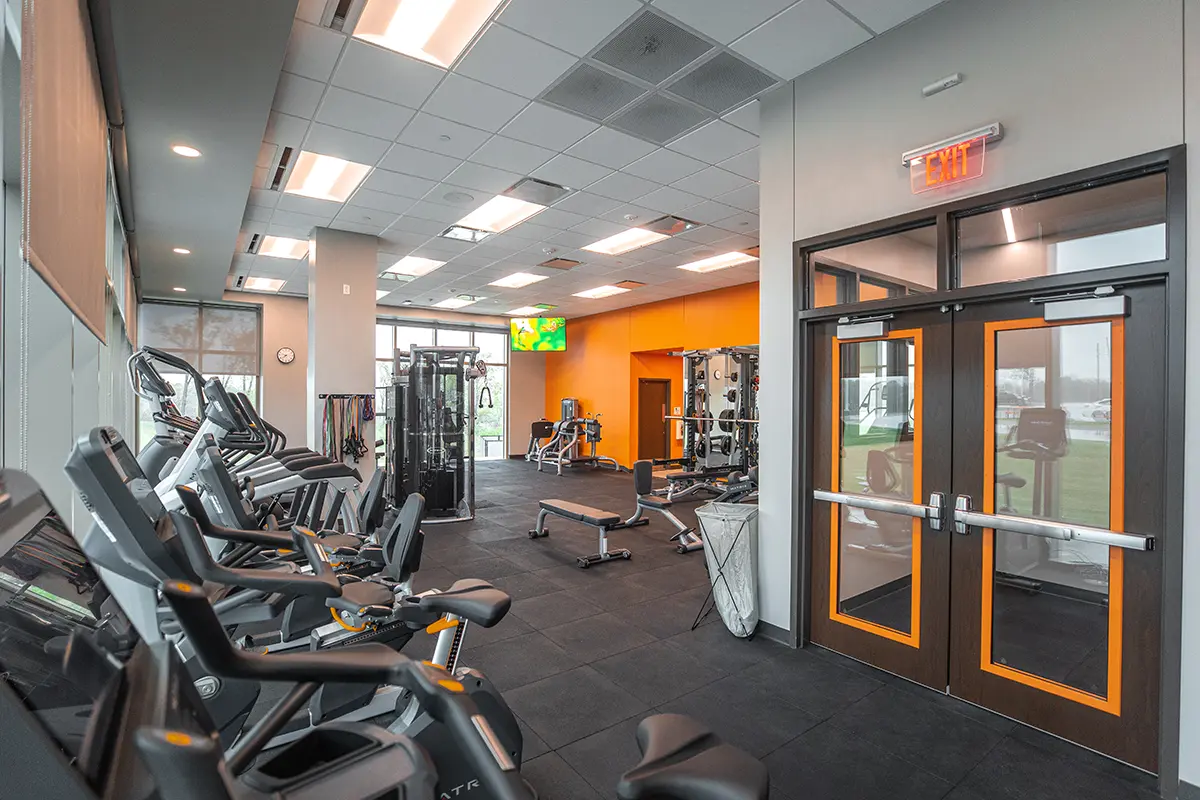 Modern gym interior with treadmills and exercise bikes on the left, strength machines on the right. Bright lighting and an orange accent wall create an energetic vibe.