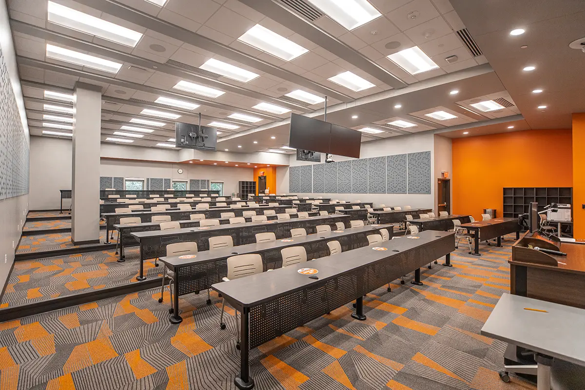 Modern lecture hall with curved rows of desks and beige chairs, featuring bright orange walls and patterned carpet. Overhead screens and bright lighting create a professional atmosphere.
