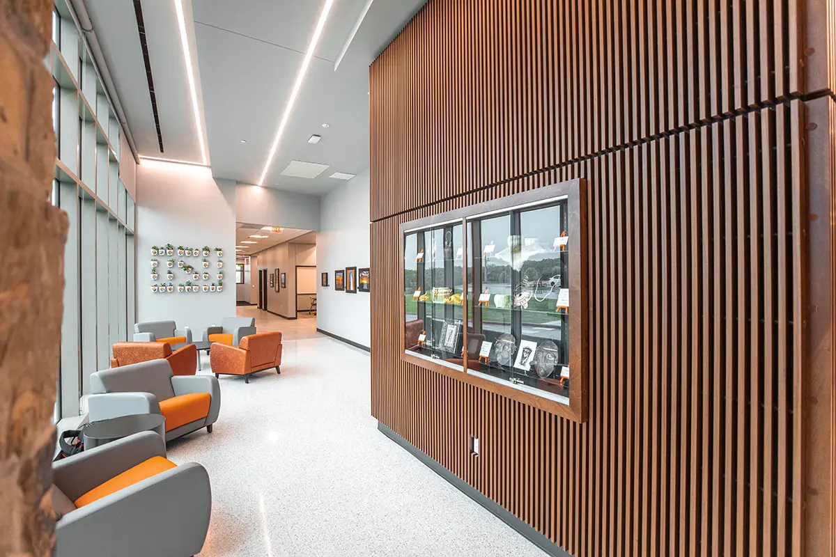 Modern lobby with wood panel walls, glass display case, and seating area with orange and gray chairs. Natural light streams through large windows.