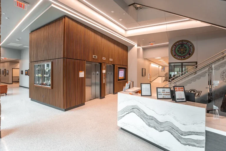 Modern lobby with a sleek marble reception desk, wood-paneled walls, and stainless steel elevators. Bright, clean lighting creates a welcoming atmosphere. A decorative emblem and signage are visible near a staircase.