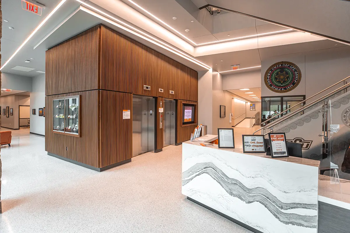 Modern lobby with a sleek marble reception desk, wood-paneled walls, and stainless steel elevators. Bright, clean lighting creates a welcoming atmosphere. A decorative emblem and signage are visible near a staircase.