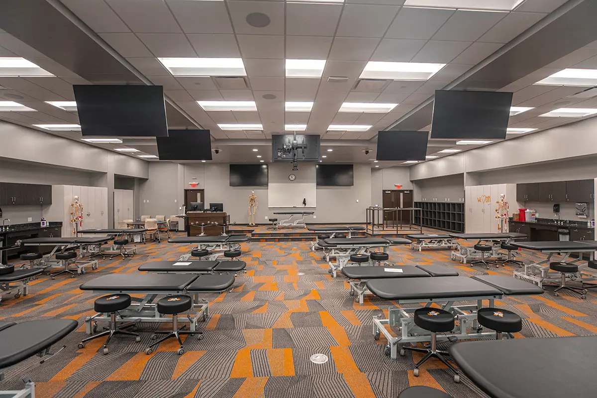 A spacious medical training room with numerous exam tables arranged neatly. Screens hang from the ceiling, and anatomical models are positioned near the walls. The room is well-lit, creating a professional and educational atmosphere.