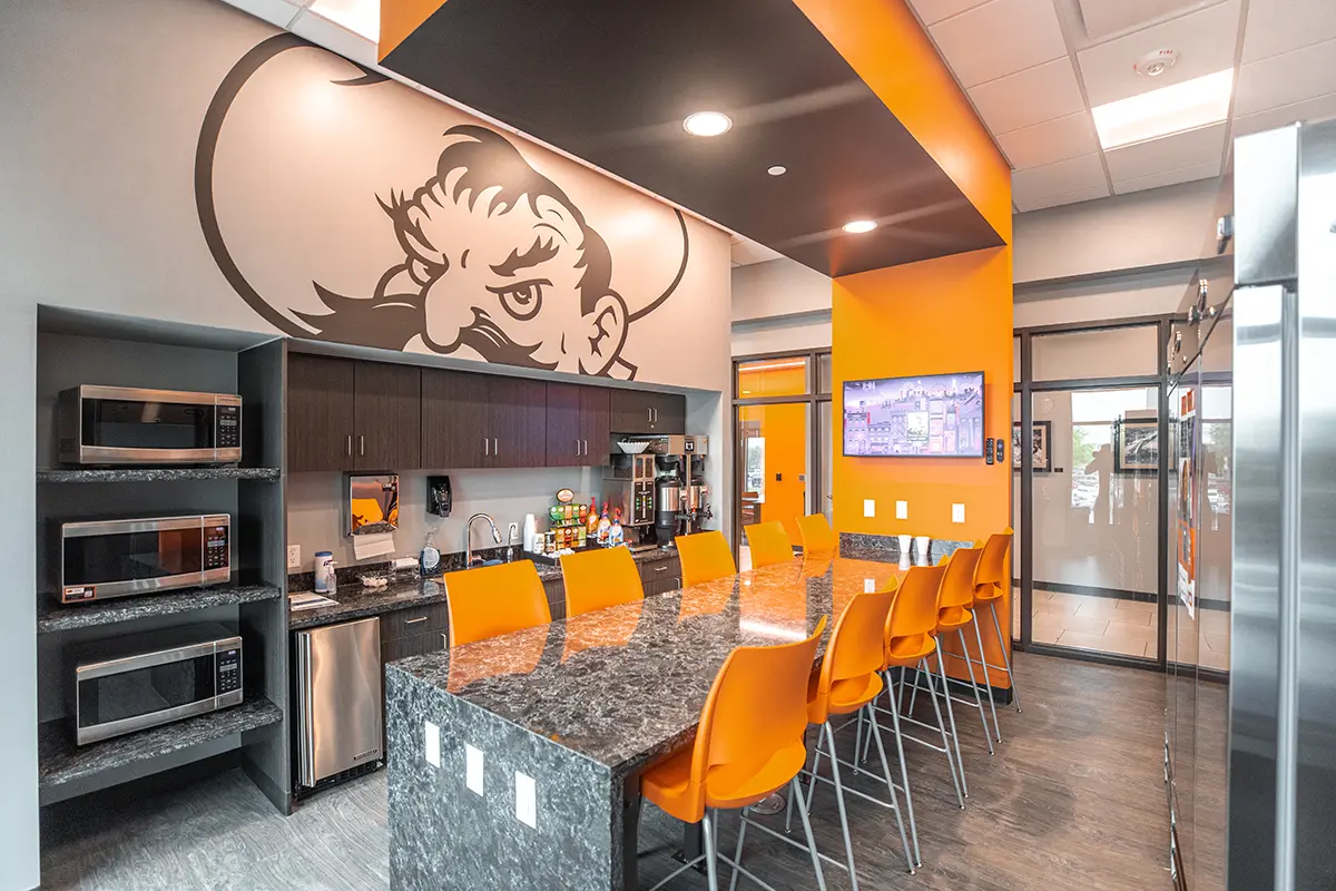 Modern break room with a stone counter surrounded by orange chairs. Left wall features three microwaves and a cowboy mural. Bright and inviting.