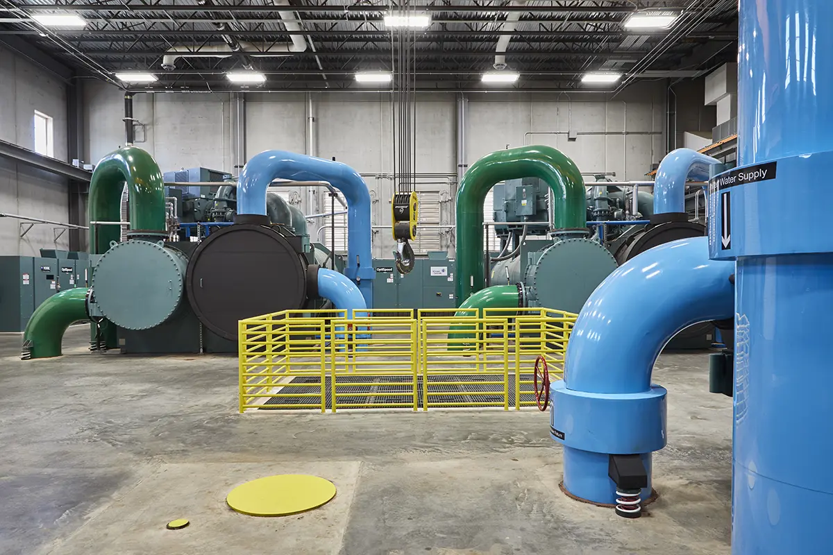 Large industrial room with blue and green pipes curving around big cylindrical tanks. A yellow metal railing and overhead lights add a clean, modern feel.