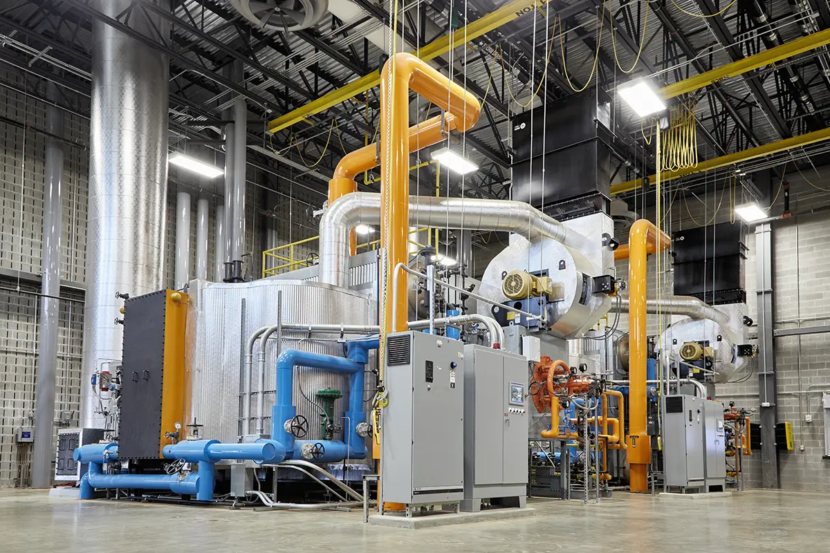 Bright industrial room with large machinery, featuring orange and blue pipes and various metal components. The atmosphere is clean and organized.