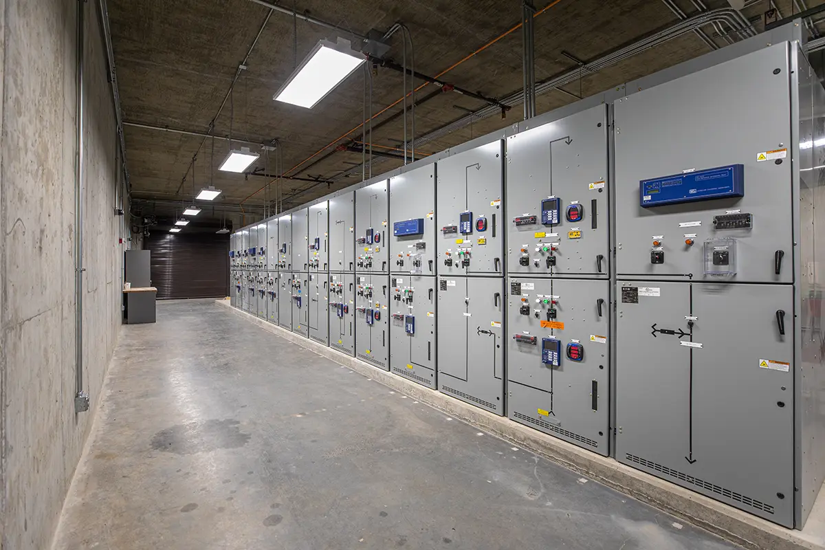 A concrete industrial room with large, gray electrical panels lined along the right wall. Bright ceiling lights illuminate the clean, organized space.