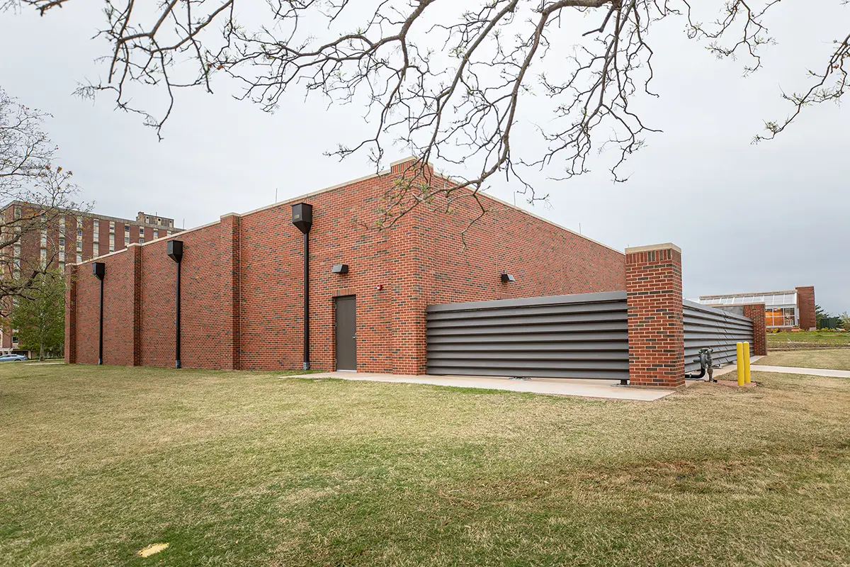 A large brick building with minimal windows stands on a grassy lawn under an overcast sky. Sparse branches frame the top. The scene feels quiet and subdued.