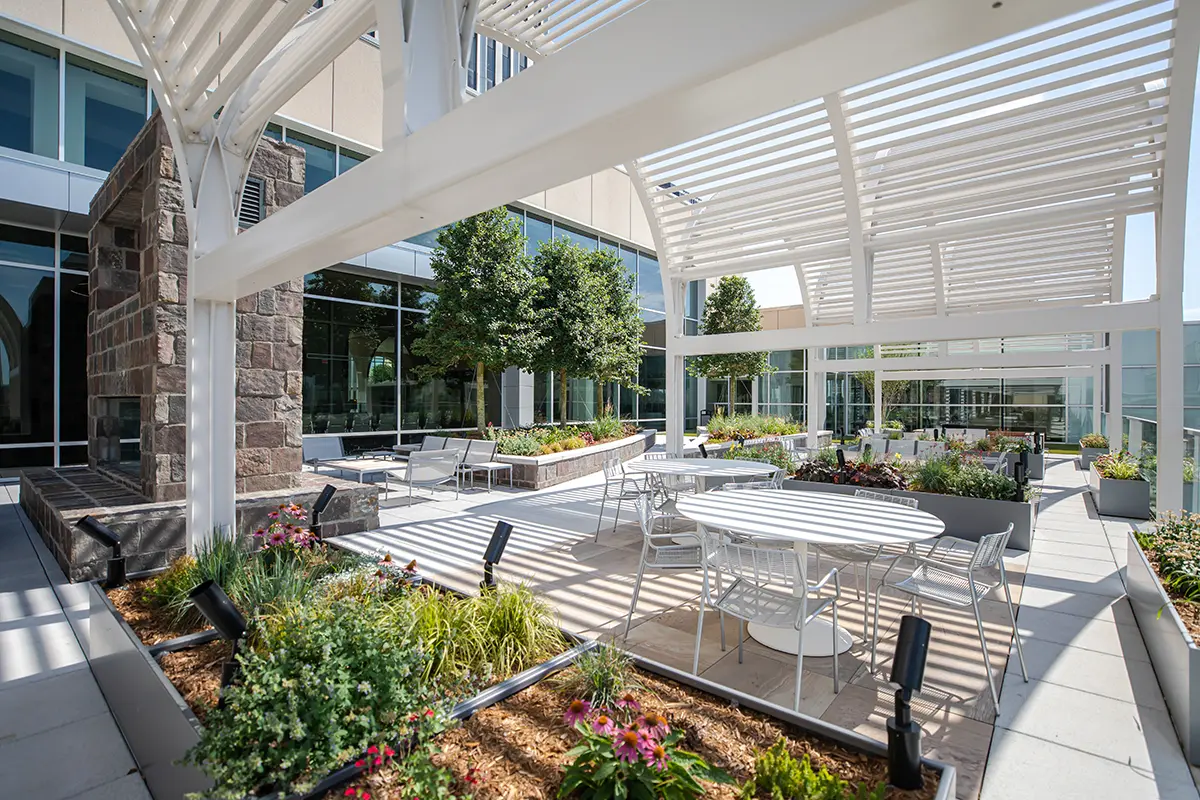 Moderno patio exterior con pérgolas blancas, mesas y sillas de metal. El espacio está rodeado de exuberante vegetación, flores de vivos colores y grandes ventanales, lo que crea un ambiente sereno y acogedor.