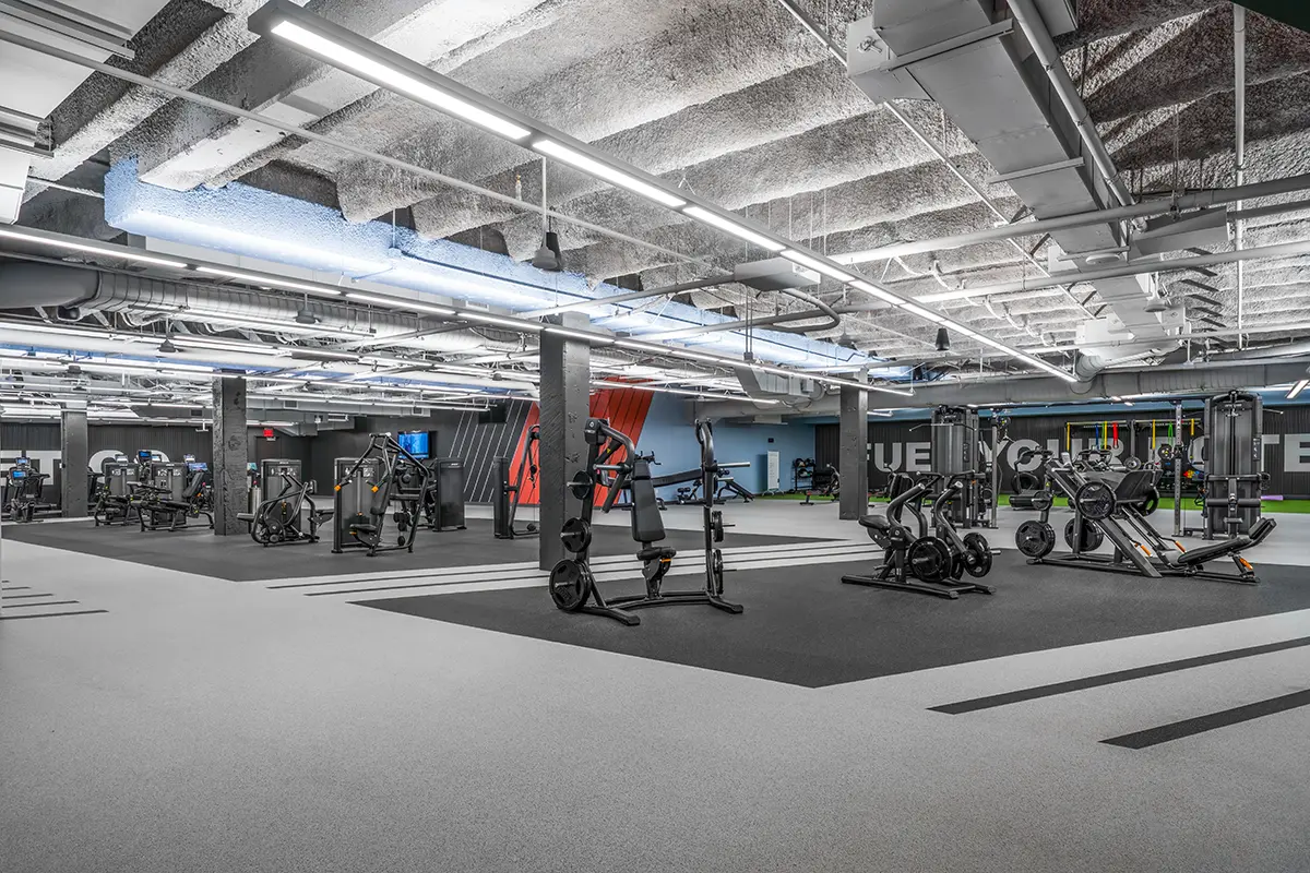 Gimnasio moderno y espacioso con varias máquinas de entrenamiento, suelo gris y negro, tuberías a la vista en el techo e iluminación brillante que crean un ambiente enérgico.