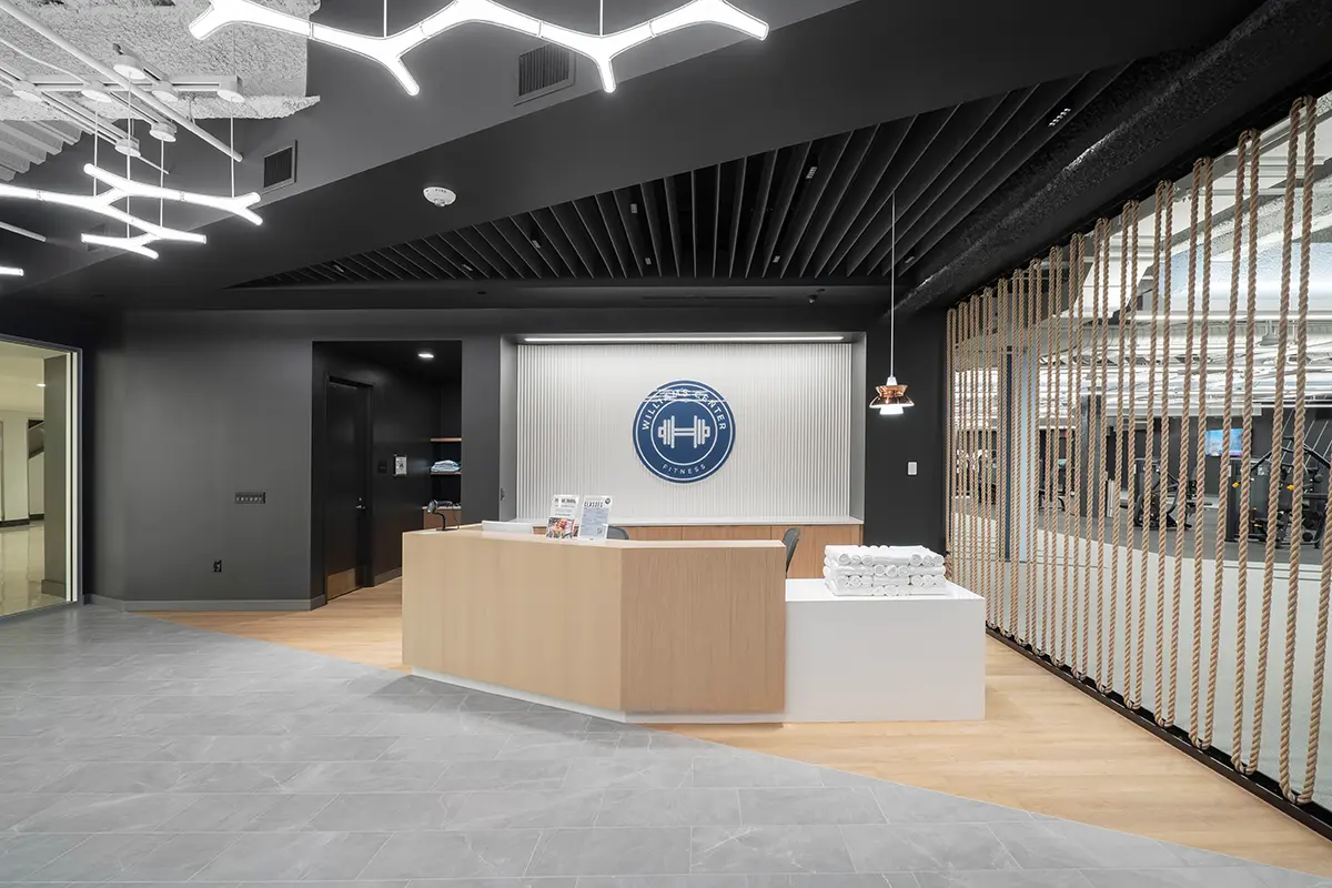 Entrada moderna al gimnasio con un elegante mostrador de recepción de madera clara, un logotipo azul sobre una pared blanca y una iluminación geométrica en el techo. Un ambiente acogedor.