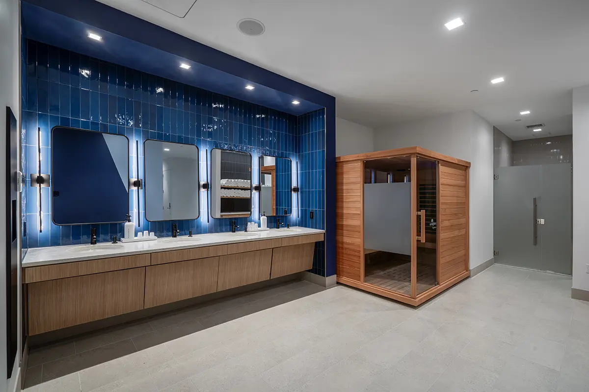 Entrada moderna al gimnasio con un elegante mostrador de recepción de madera clara, un logotipo azul sobre una pared blanca y una iluminación geométrica en el techo. Un ambiente acogedor.