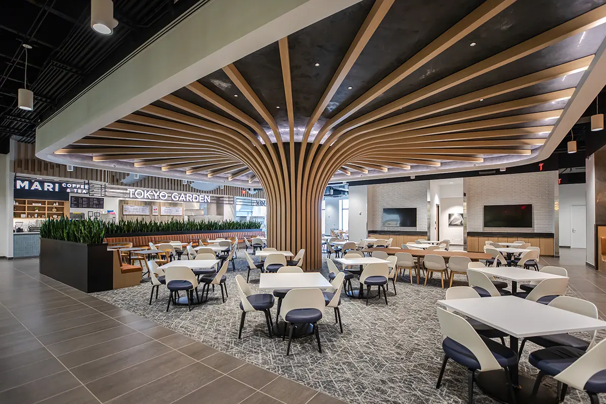 Amplio y moderno comedor con vigas geométricas en el techo, mesas y sillas elegantes. Ambiente luminoso y aireado con carteles de «Tokyo Garden» y cafetería.