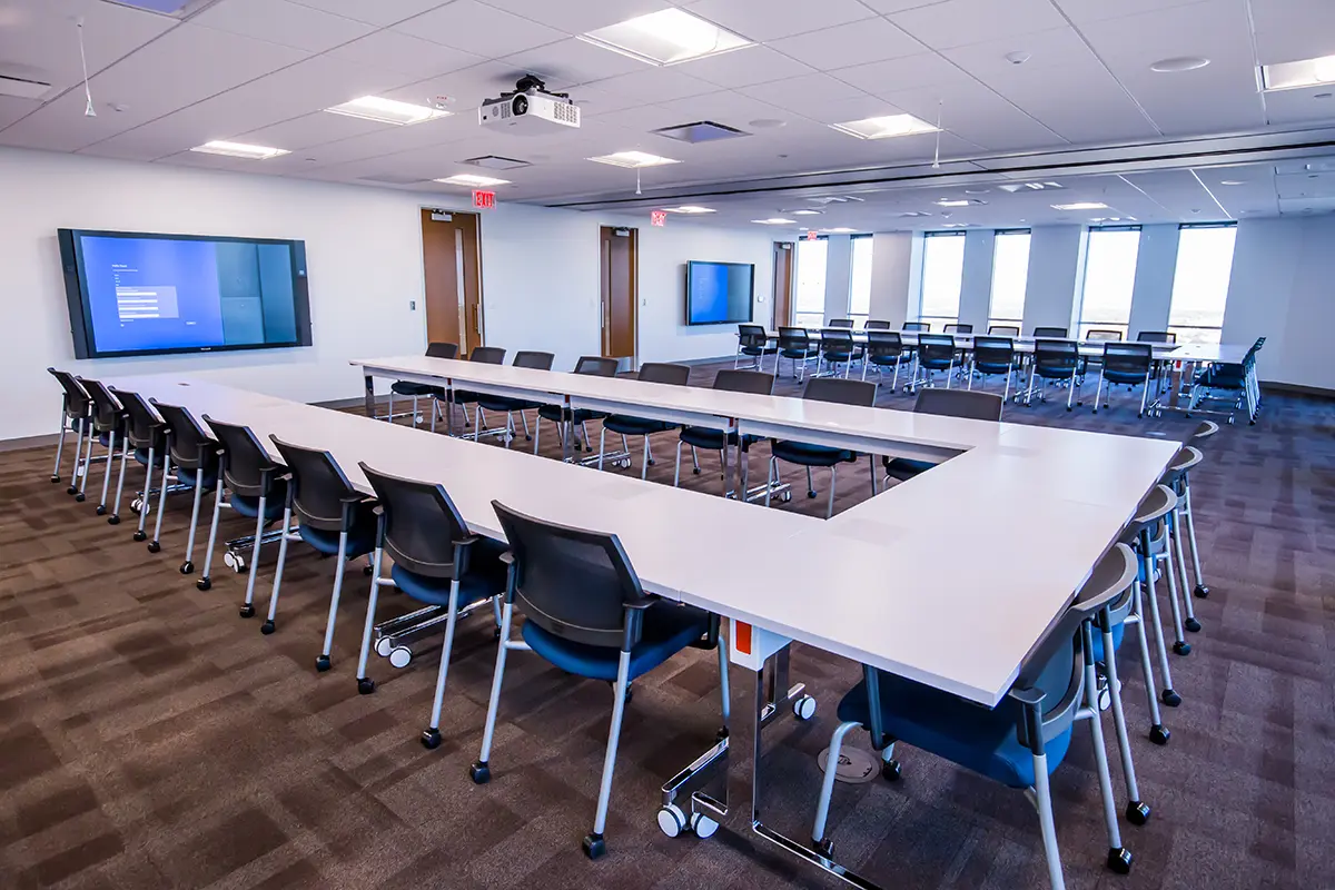 Amplia sala de conferencias con mesas dispuestas en forma de U, sillas modernas y dos pantallas montadas en la pared. La iluminación brillante y los tonos neutros crean un ambiente profesional.