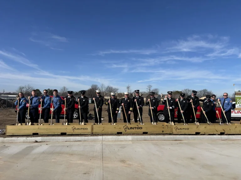 A group of people in uniforms and business attire hold shovels at a groundbreaking ceremony. They stand on a platform with "FLINTCO" branding under a clear blue sky.