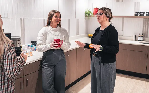 Two women in an office kitchen, chatting and smiling. One holds a red mug, and the other holds an orange. The atmosphere is casual and friendly.