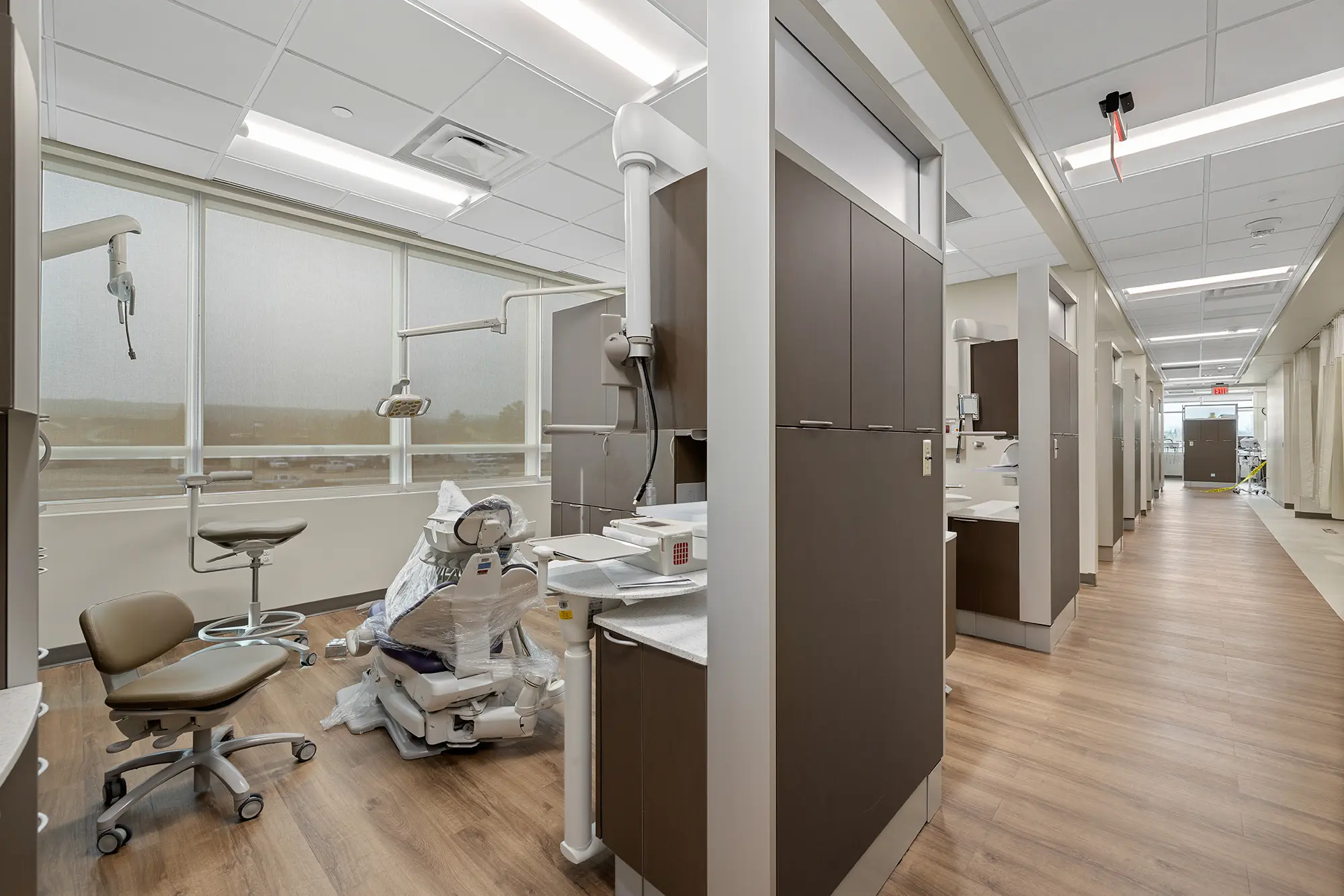 A modern dental clinic with wooden floors, dental chair, and equipment. Bright natural light streams through large windows, creating a clean, sterile atmosphere.
