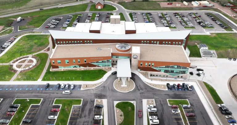Aerial view of a large, modern brick building with a white roof and expansive parking lots. Green lawns surround the structure, evoking a sense of openness.