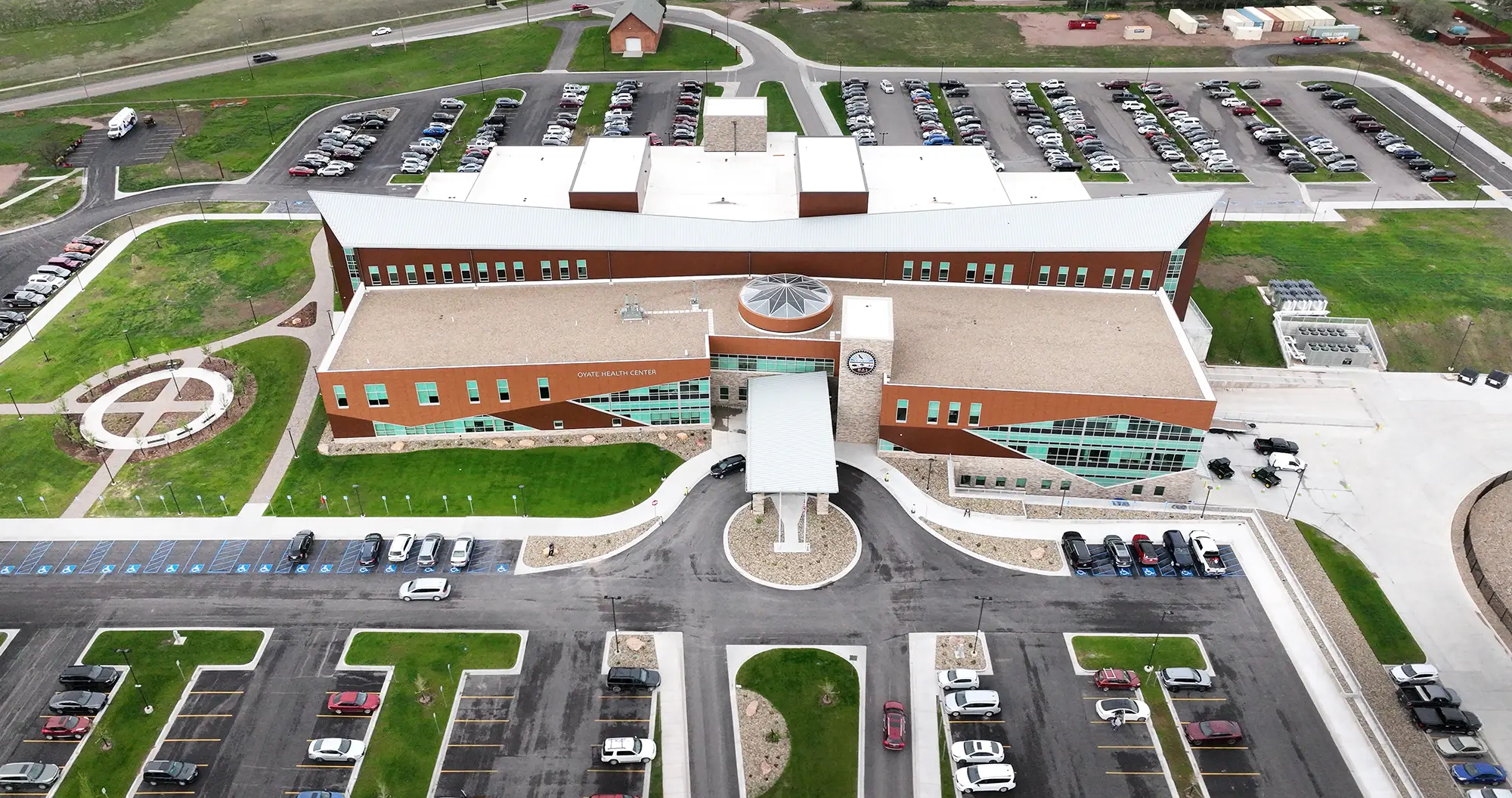 Aerial view of a large, modern brick building with a white roof and expansive parking lots. Green lawns surround the structure, evoking a sense of openness.