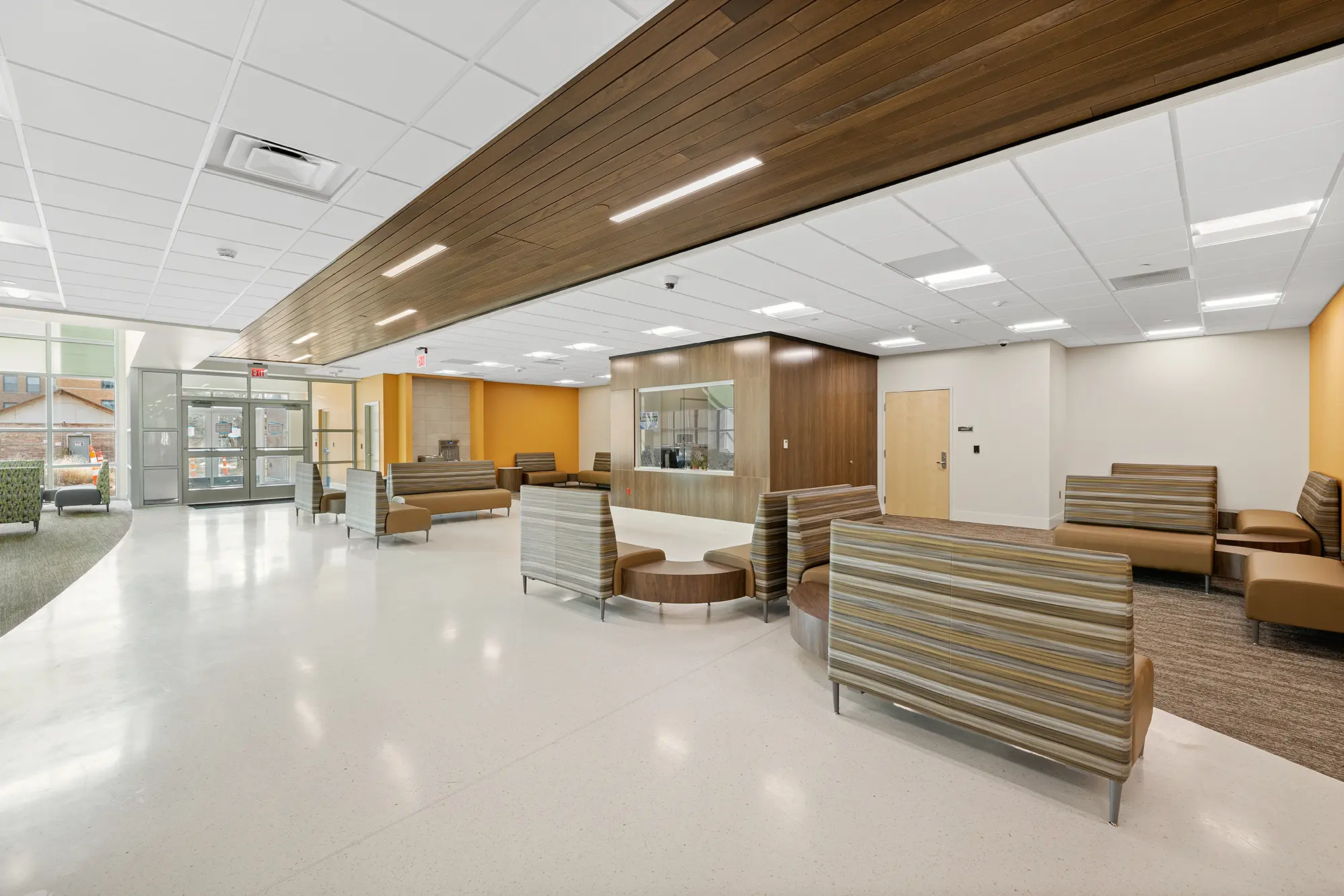 Modern, spacious waiting area with neutral tones. Features striped couches, orange accent walls, wood panel ceiling, and large windows for natural light.