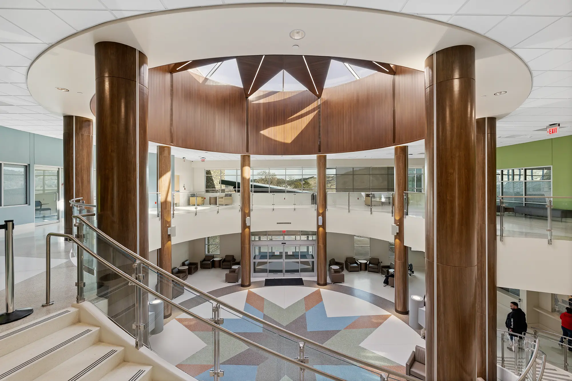 Spacious lobby with geometric tile floor, surrounded by sleek wooden columns and a circular skylight. Modern, airy atmosphere with soft natural light.