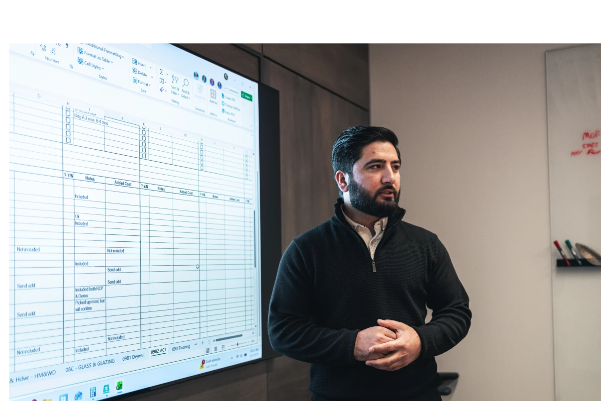 A man in a dark sweater presents data from a spreadsheet on a large screen. He appears focused and engaged, standing in a modern office setting.