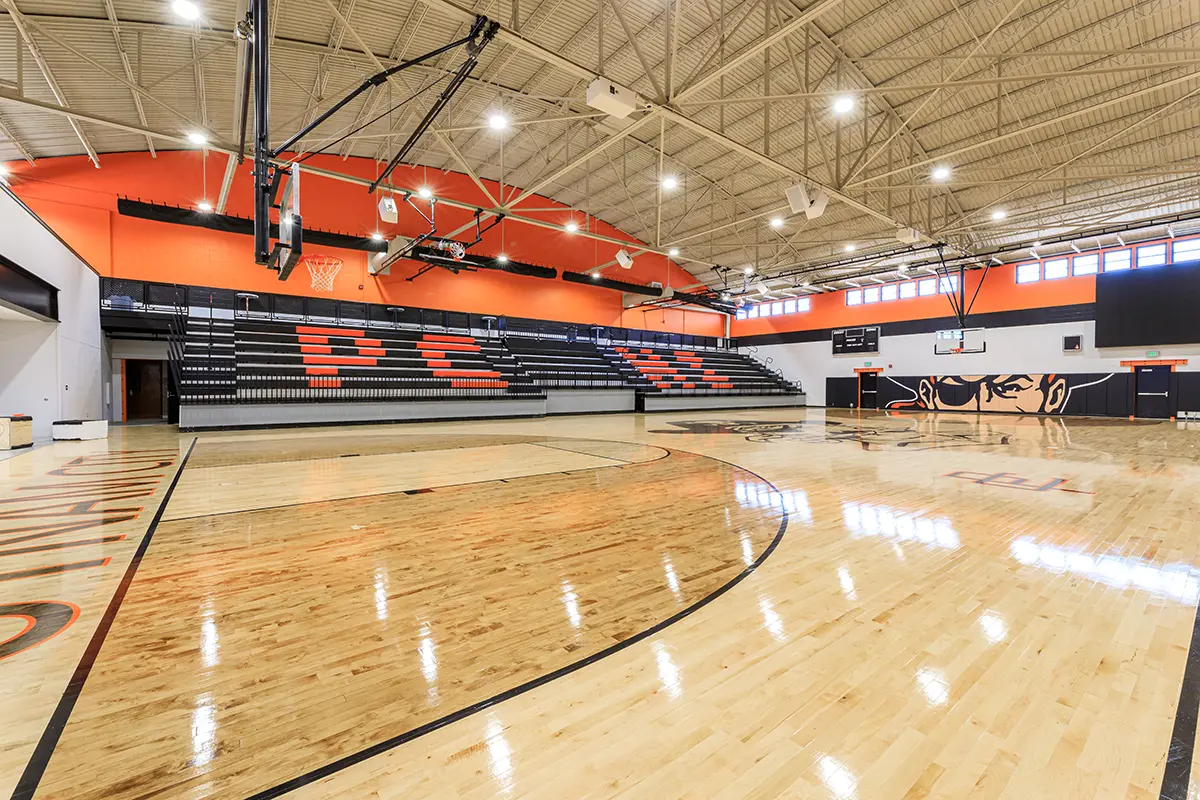Bright gymnasium with polished wooden floors, high ceilings, and orange accents. Empty bleachers display "PC" in orange seats. Quiet, spacious atmosphere.