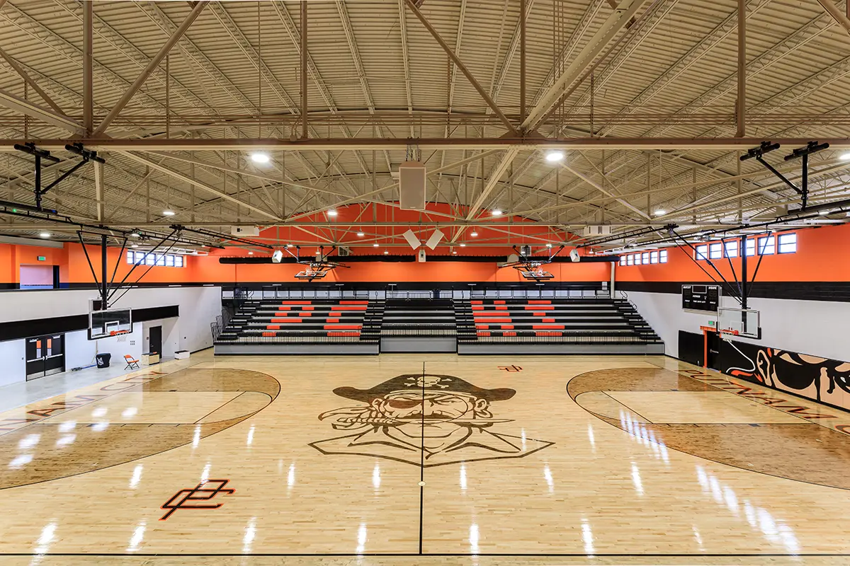 Spacious high school gym with polished wooden floor, featuring a large pirate logo at center court. "PC HI" is emblazoned on the orange-black bleachers.