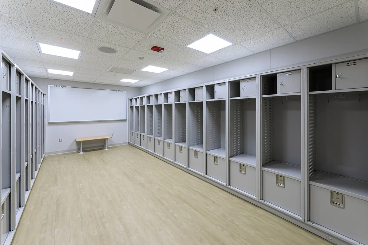 A spacious, empty locker room with gray metal lockers lining two walls. It features a light wood floor and bright overhead lighting, creating a clean, sterile ambiance.