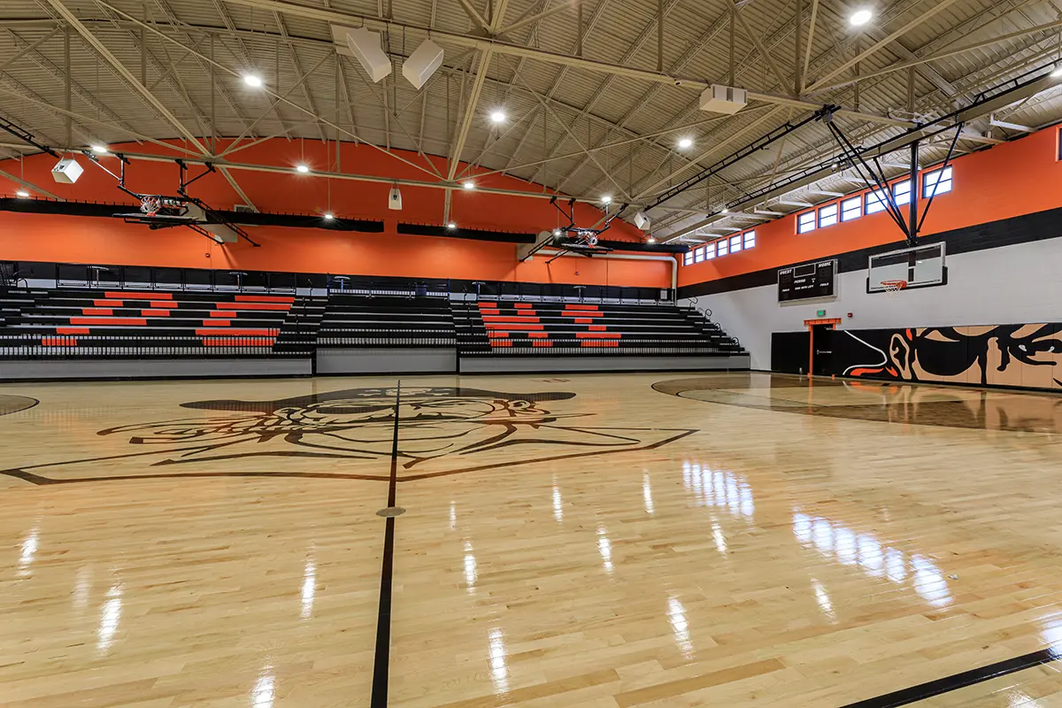 A brightly lit indoor basketball court with polished wooden floors featuring a large mascot design at center court. Orange and black bleachers spell "PCH" at the far end, creating a vibrant and energetic atmosphere.